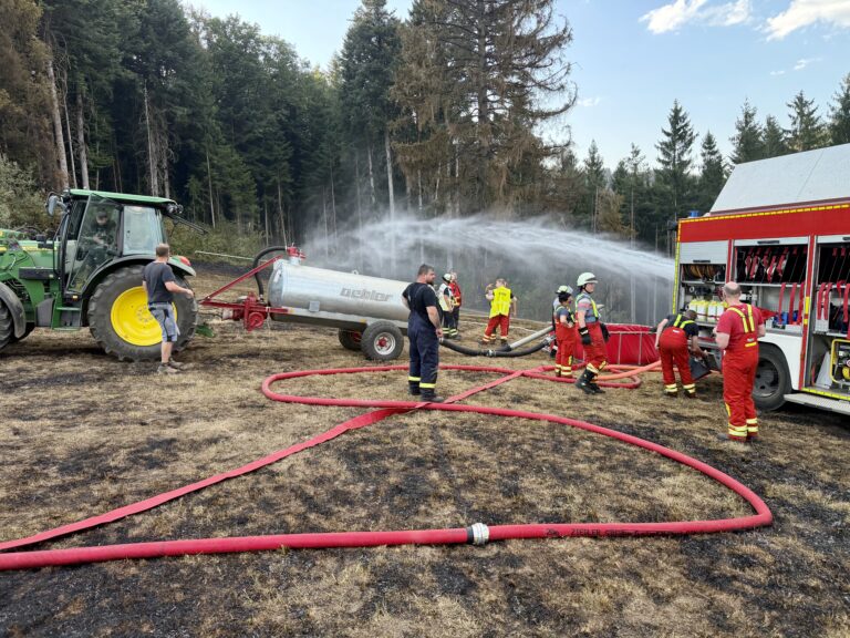 Firefighters connect hoses to a water tanker in a forest clearing; a red fire truck is visible on the right side.