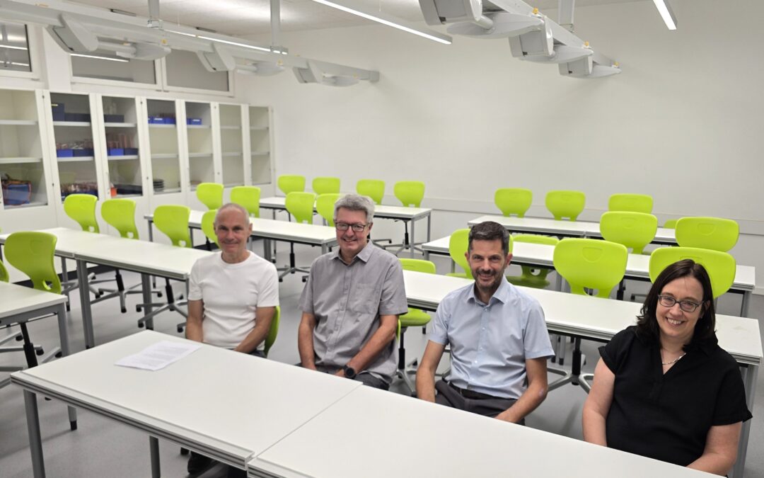 Four adults sit at white desks in a bright classroom with lime-green chairs and white storage cabinets along the back wall.