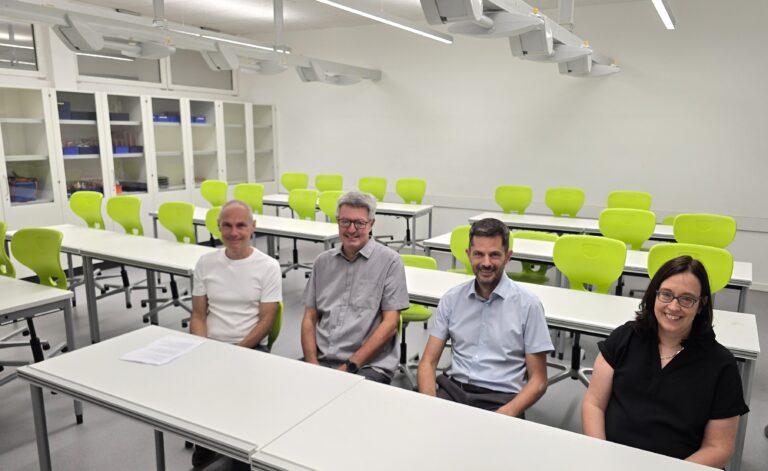 Four adults sit at white desks in a bright classroom with lime-green chairs and white storage cabinets along the back wall.