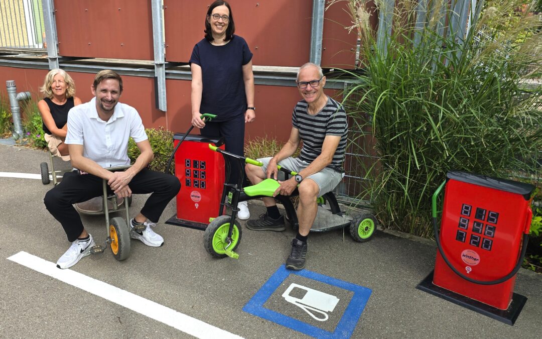 Four adults posing with pedal-assisted mobility trikes in a parking lot, smiling in front of a red wall and greenery.