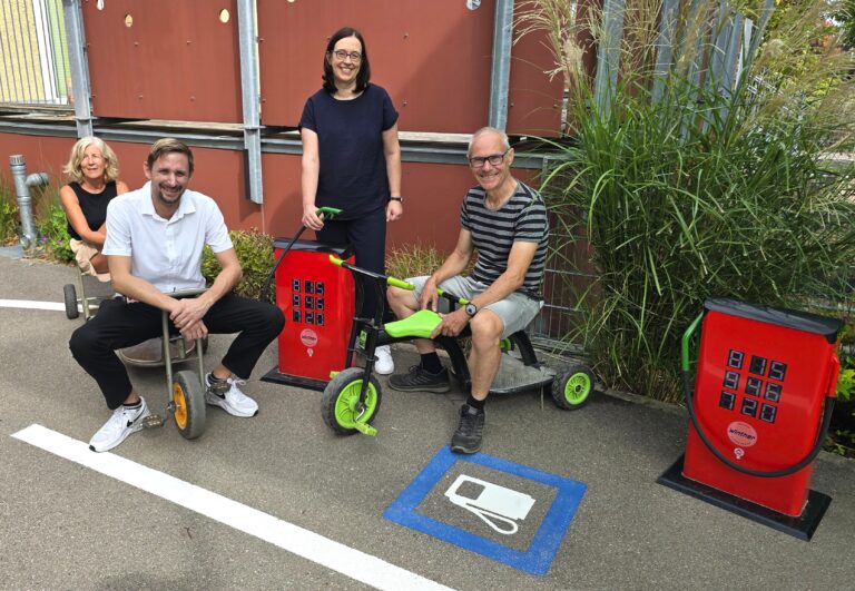 Four adults posing with pedal-assisted mobility trikes in a parking lot, smiling in front of a red wall and greenery.