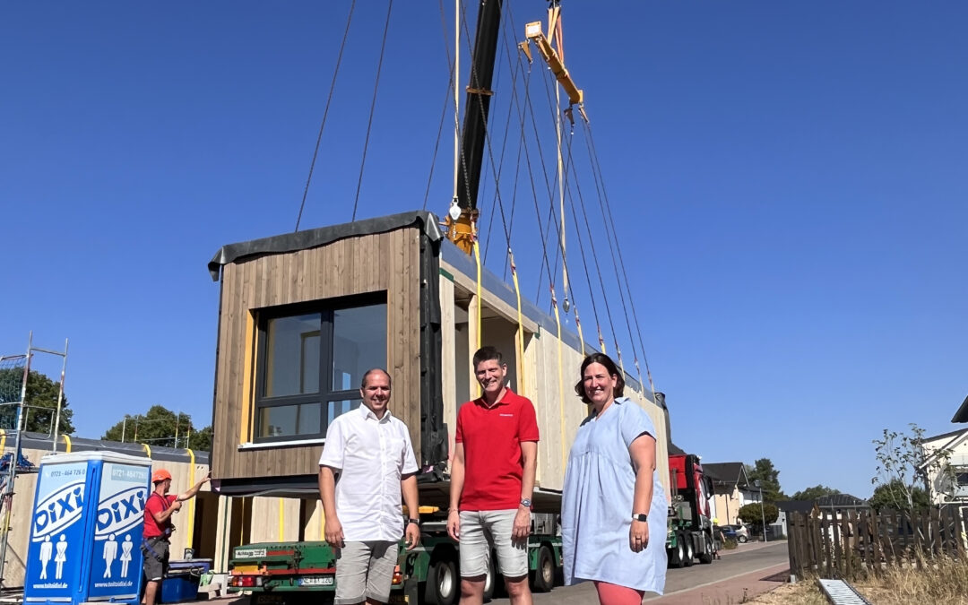 Three adults stand on a sunny street in front of a partially built modular building being lifted by a crane on a truck, clear blue sky above.