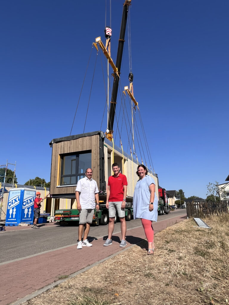 Three adults stand on a sunny street in front of a partially built modular building being lifted by a crane on a truck, clear blue sky above.