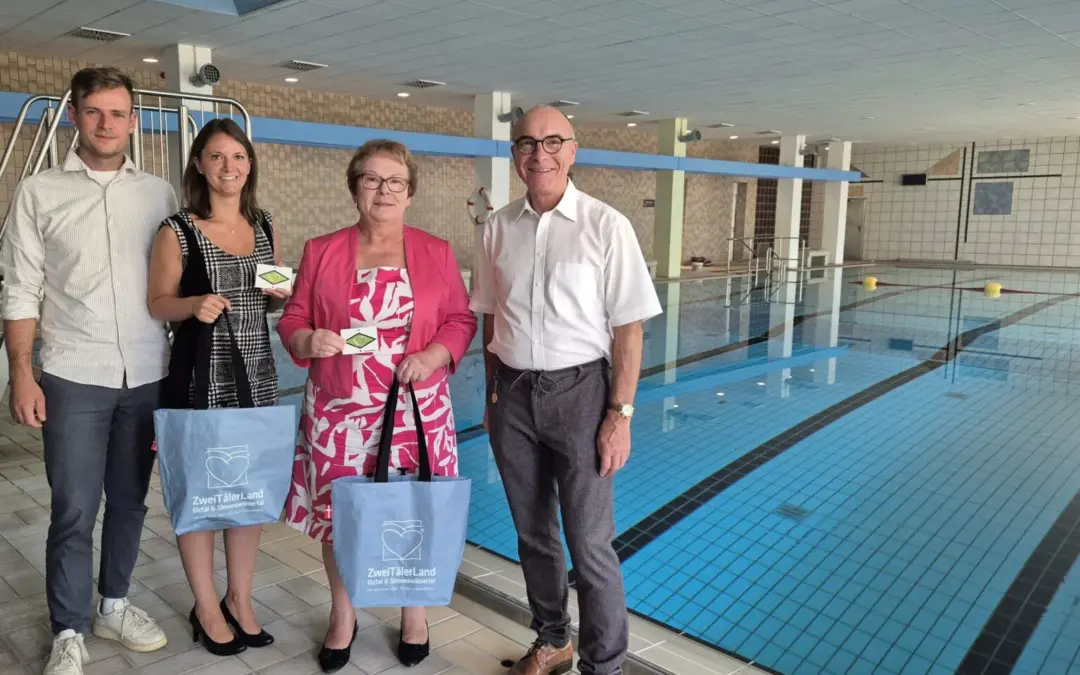 Four adults pose by an indoor swimming pool, two women hold blue tote bags and small cards, smiling at the camera.