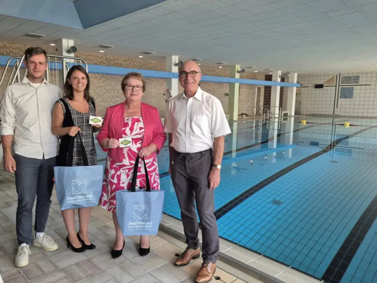 Four adults pose by an indoor swimming pool, two women hold blue tote bags and small cards, smiling at the camera.