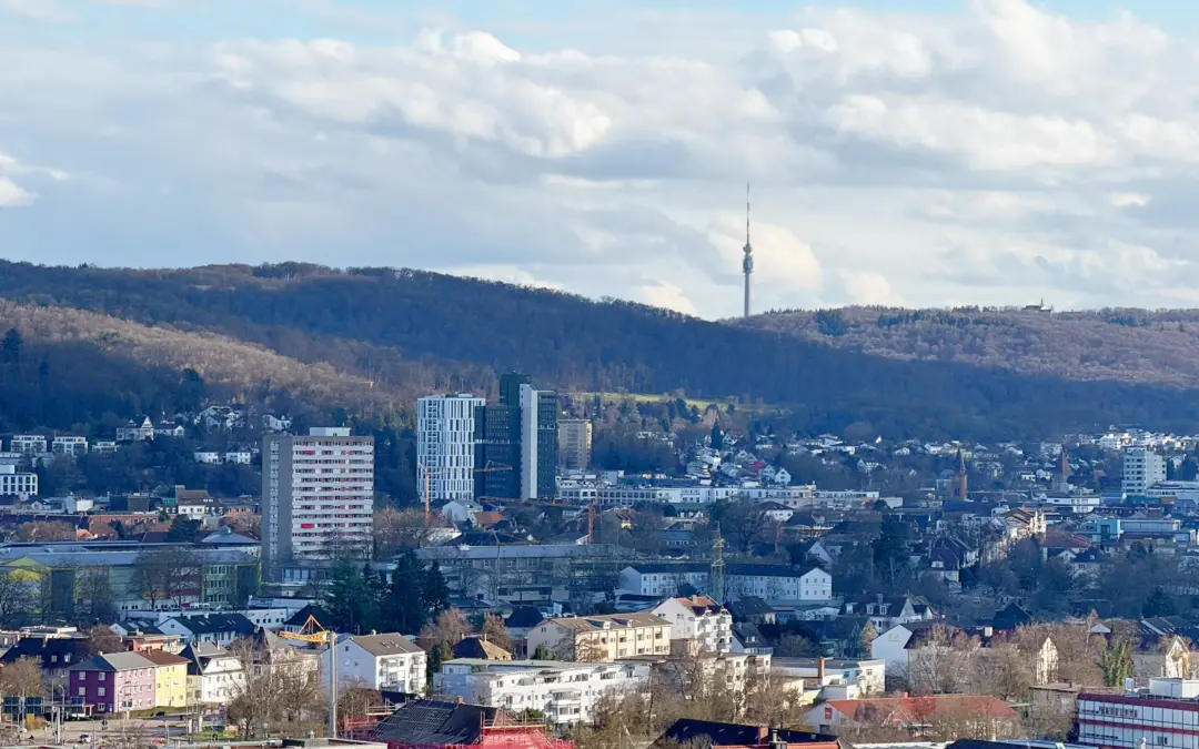 City skyline with a tall antenna tower on a hill, surrounded by low-rise buildings and forested hills under a blue sky with clouds.
