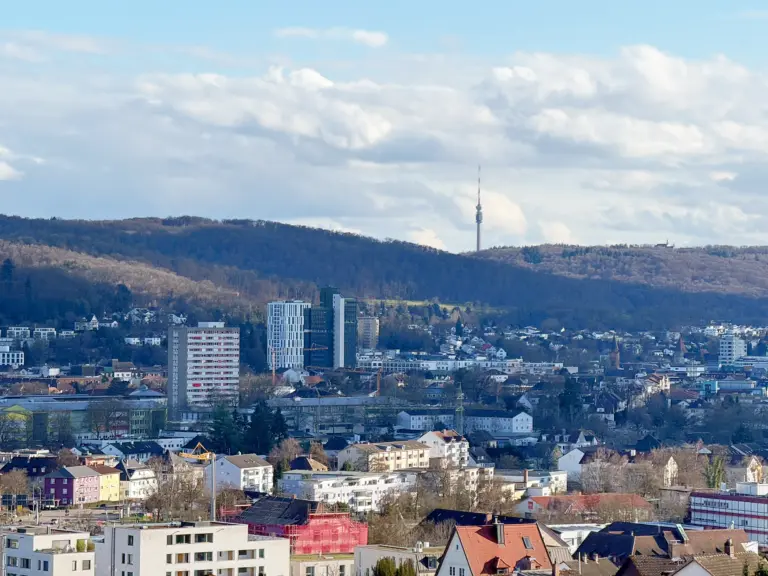 City skyline with a tall antenna tower on a hill, surrounded by low-rise buildings and forested hills under a blue sky with clouds.