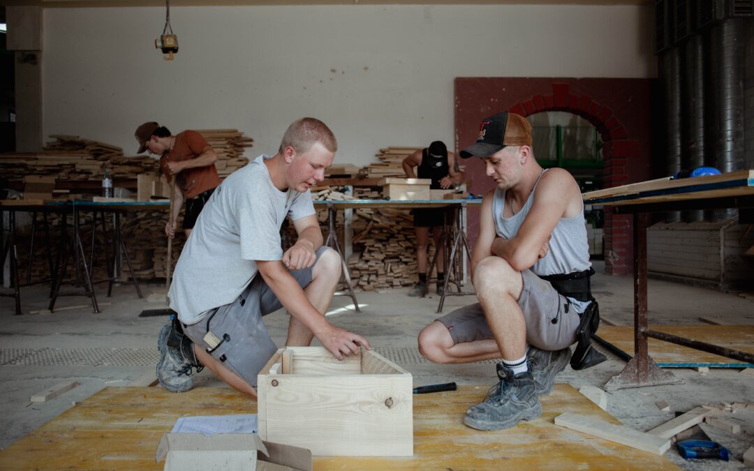 Three workers in a woodshop assemble a wooden crate, with stacks of lumber and tools in the background.