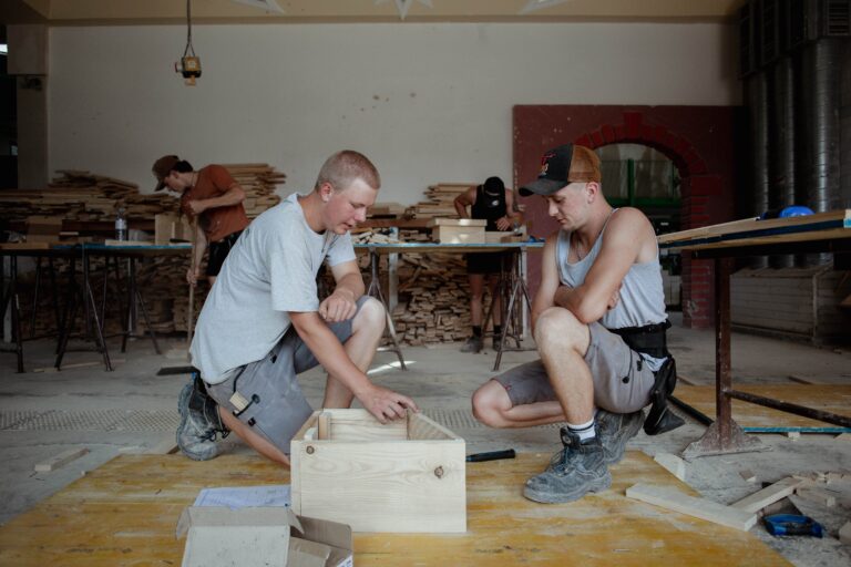Three workers in a woodshop assemble a wooden crate, with stacks of lumber and tools in the background.