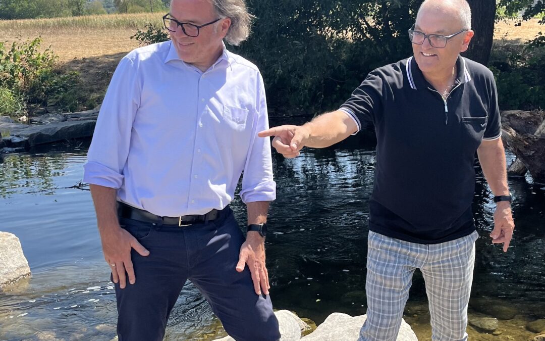 Two middle-aged men stand on a rocky lakeshore, one in a white shirt and dark pants and the other in a black polo and plaid pants, with the man in black pointing off to the side by a calm pond and a large tree behind them.