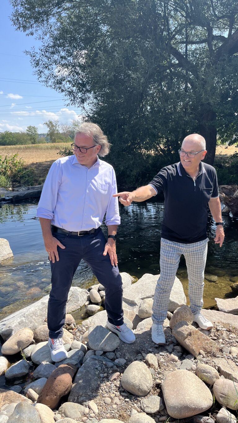 Two middle-aged men stand on a rocky lakeshore, one in a white shirt and dark pants and the other in a black polo and plaid pants, with the man in black pointing off to the side by a calm pond and a large tree behind them.