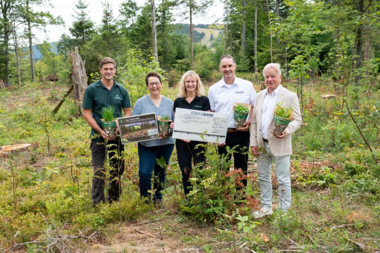 Five adults stand in a forest clearing, smiling and holding potted plants and a large donation check for a reforestation project.