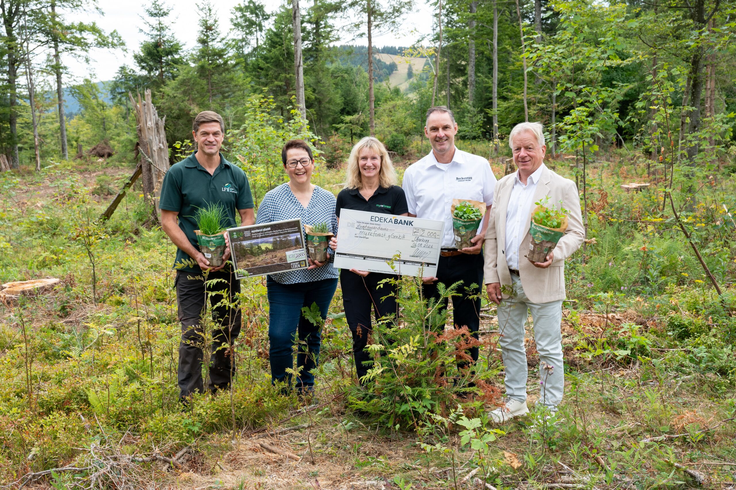 Five adults stand in a forest clearing, smiling and holding potted plants and a large donation check for a reforestation project.