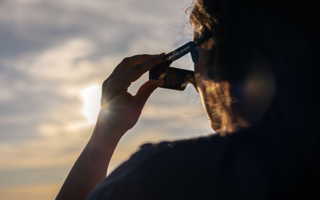Person wearing solar eclipse glasses up to the eye, with a sunset sky in the background and soft backlight.