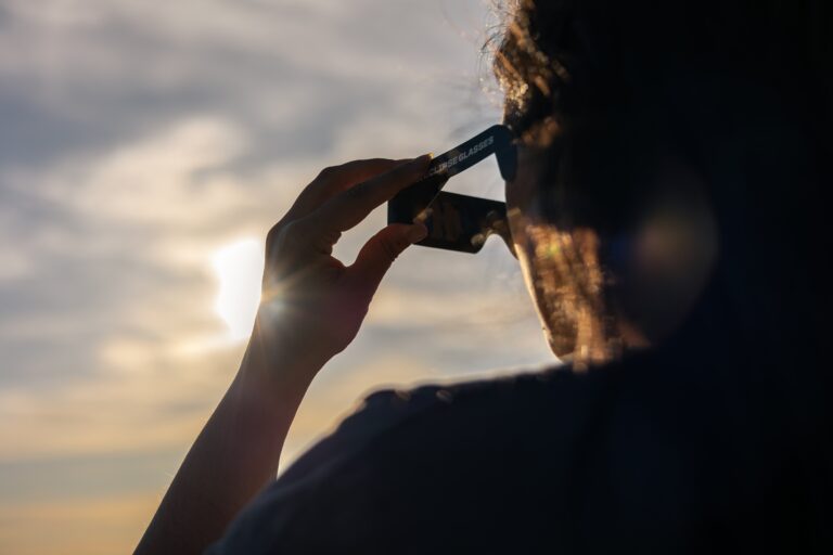 Person wearing solar eclipse glasses up to the eye, with a sunset sky in the background and soft backlight.