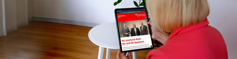Person in a red jacket holds a tablet showing a Sparkasse advertisement with three men in suits on a red banner at the top.