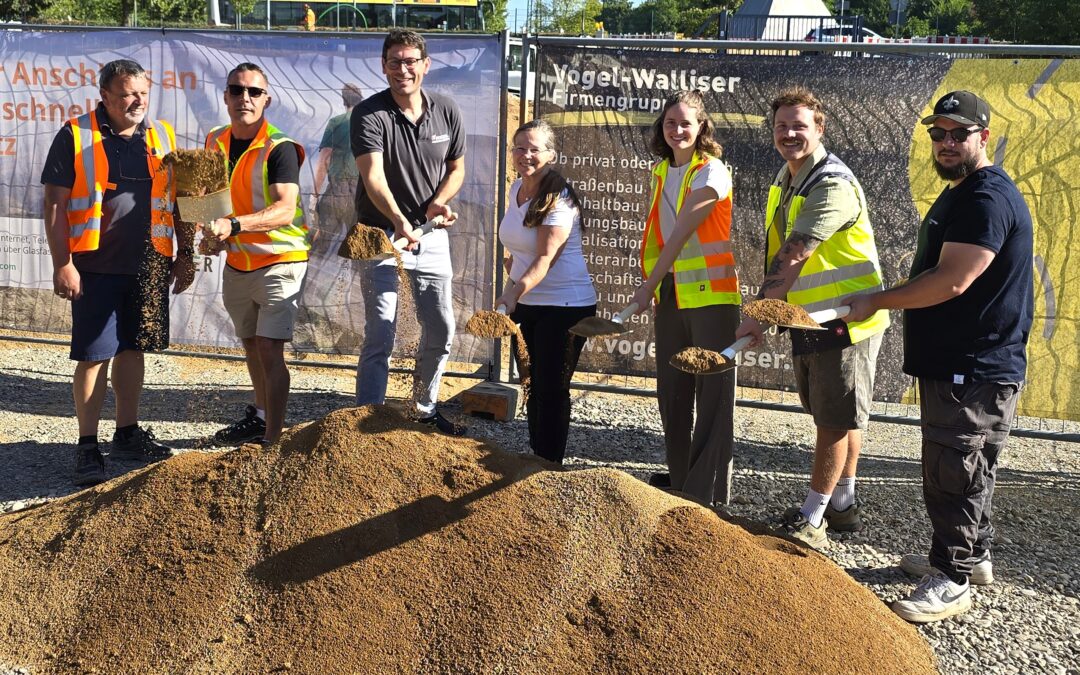 Group of construction workers in safety vests and casual clothes, smiling, holding shovels and standing beside dirt piles at a site fence backdrop.
