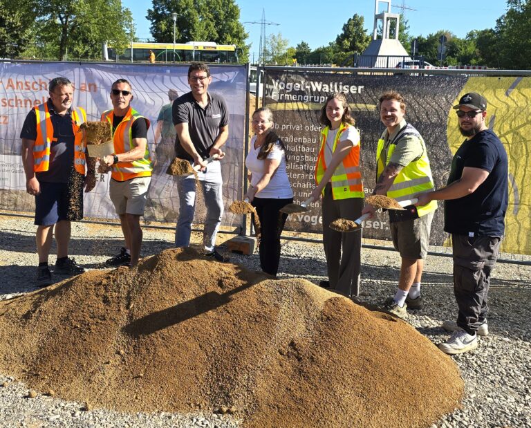 Group of construction workers in safety vests and casual clothes, smiling, holding shovels and standing beside dirt piles at a site fence backdrop.