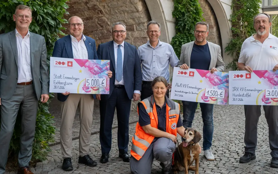 Six adults stand outdoors, holding oversized donation checks while a woman in an orange safety vest sits with a dog in front.