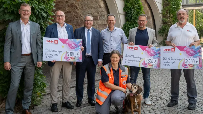 Six adults stand outdoors, holding oversized donation checks while a woman in an orange safety vest sits with a dog in front.