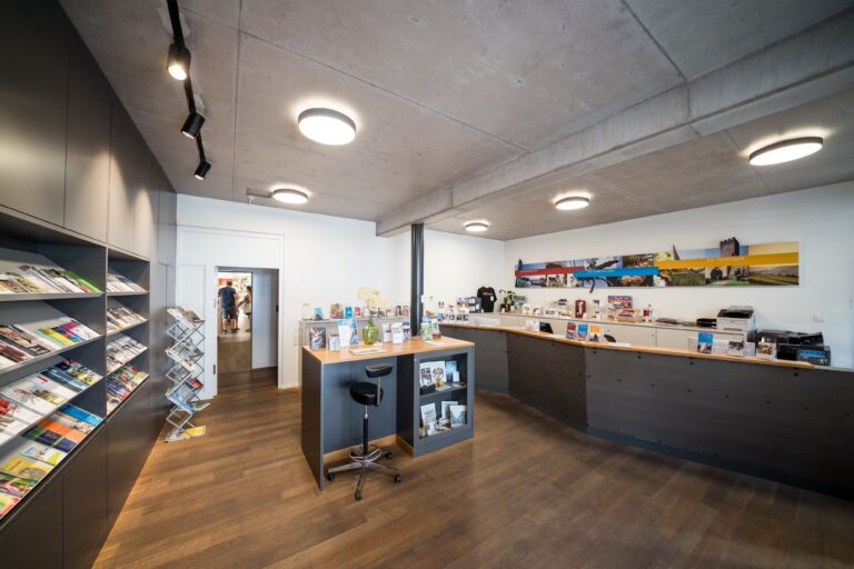 Open-plan store interior with magazine racks along the left wall and a central reception counter under round ceiling lights.