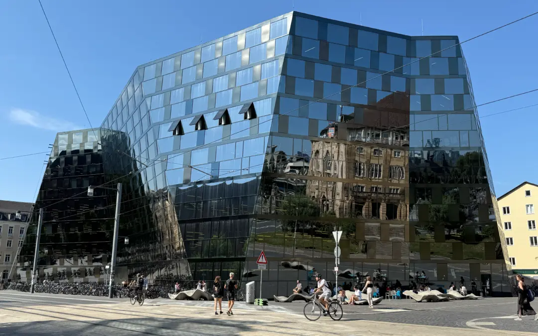 Modern glass office building with angular facade reflecting the blue sky, tram wires crisscrossing the foreground, and pedestrians and cyclists at street level.