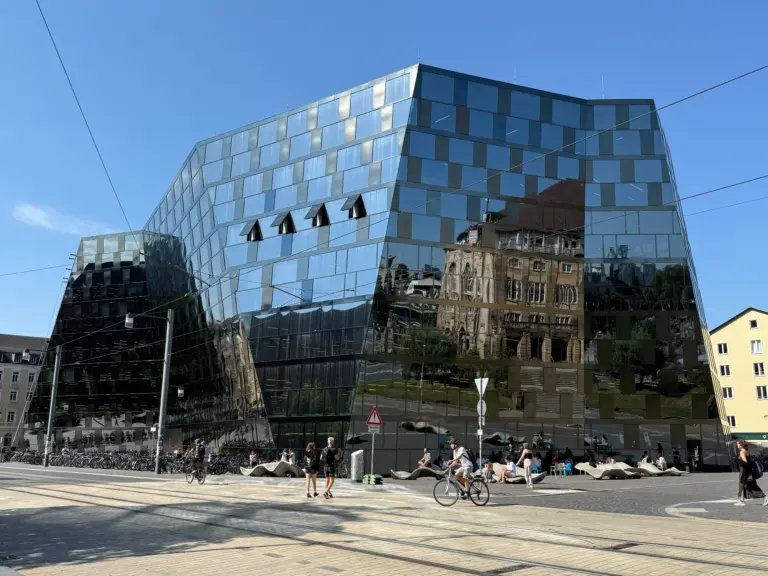 Modern glass office building with angular facade reflecting the blue sky, tram wires crisscrossing the foreground, and pedestrians and cyclists at street level.