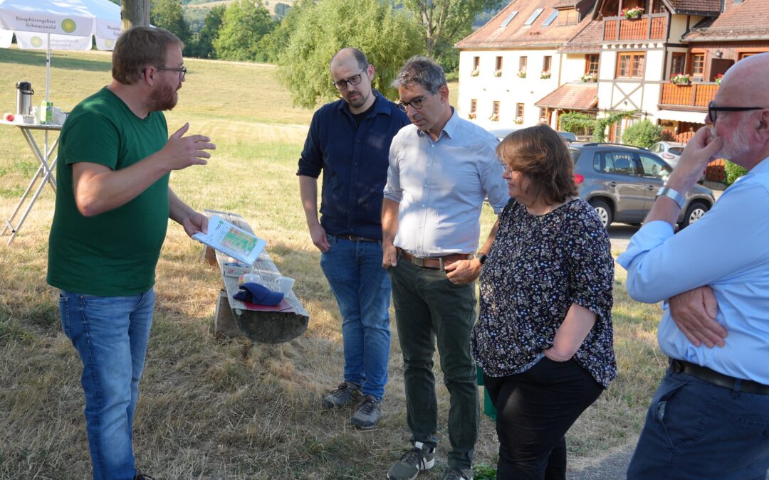 Man in green shirt explains a map to three adults outdoors in a rural setting with a white umbrella and wooden house in the background