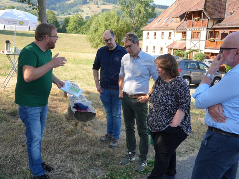 Man in green shirt explains a map to three adults outdoors in a rural setting with a white umbrella and wooden house in the background