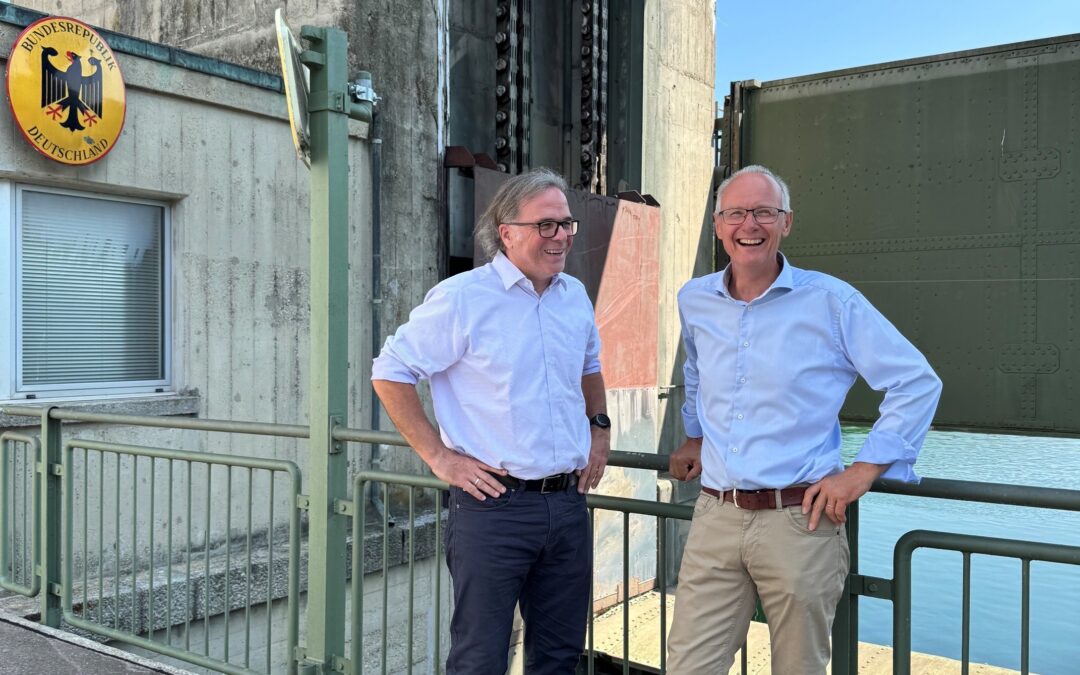 Two men in light blue shirts stand at a railing beside a concrete industrial structure, smiling at the camera with water to the right and a German sign visible on the wall behind them