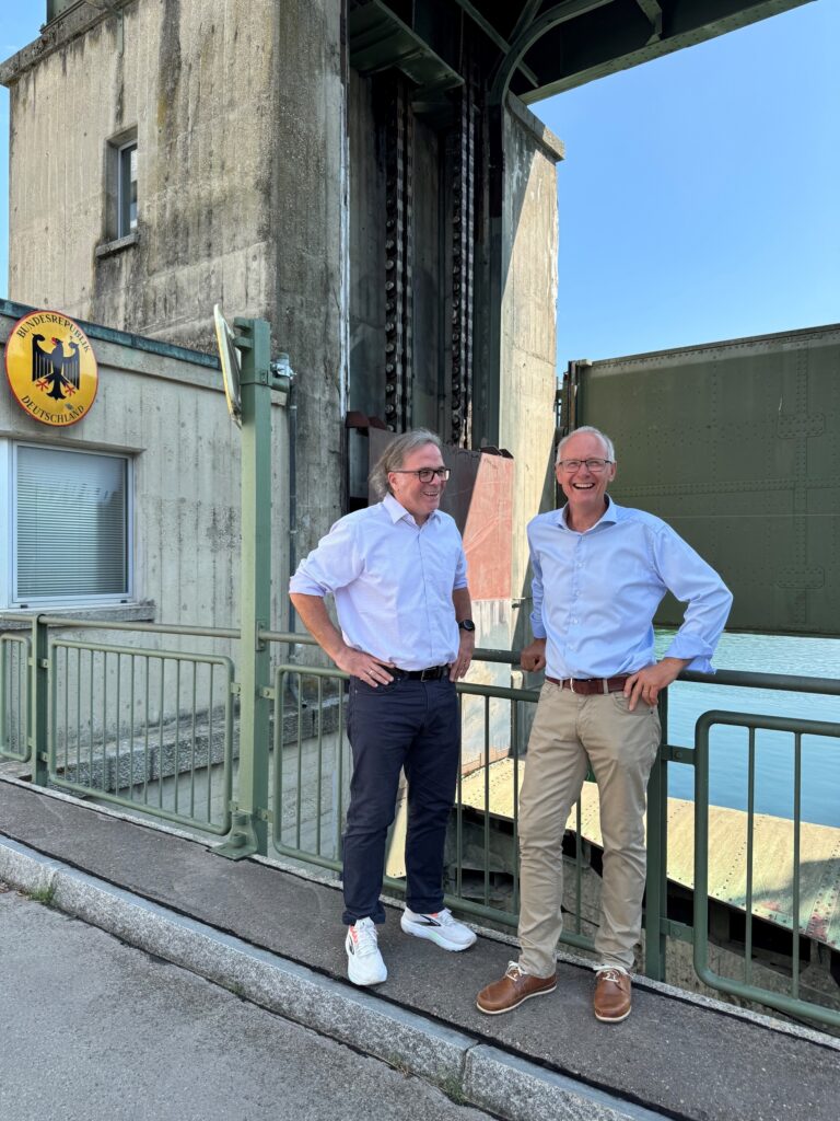 Two men in light blue shirts stand at a railing beside a concrete industrial structure, smiling at the camera with water to the right and a German sign visible on the wall behind them