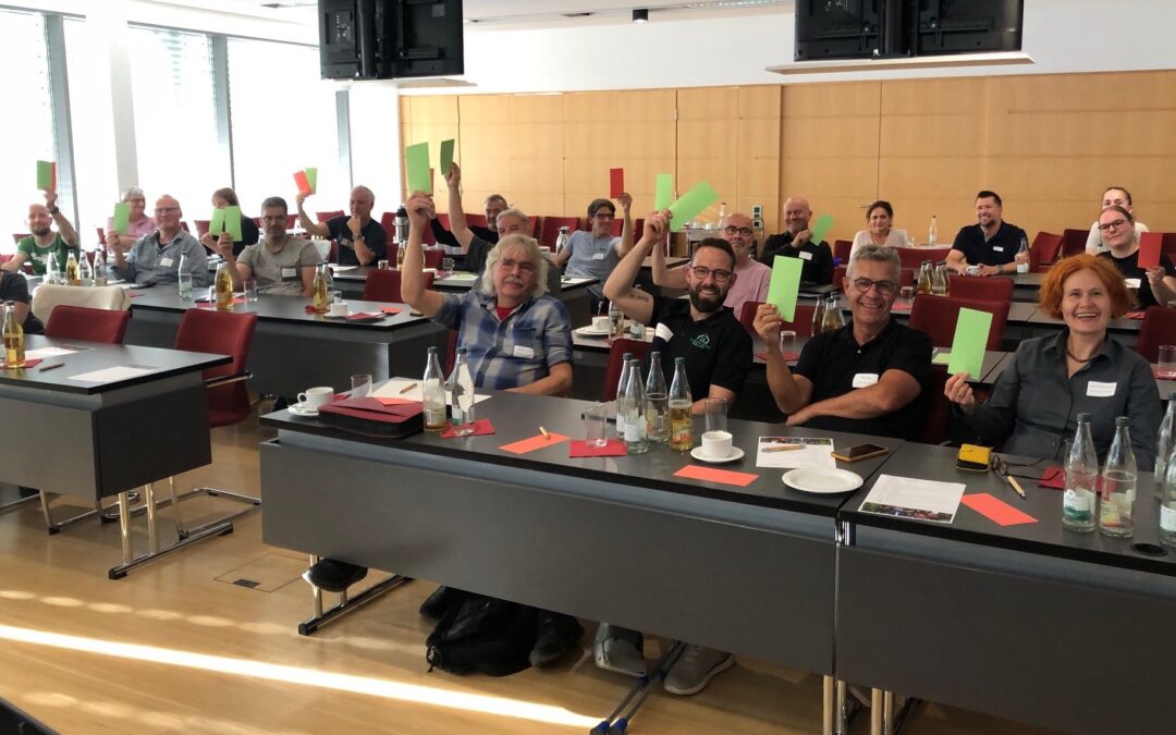 Group of adults in a classroom-style conference room, many holding up green and red cards during a discussion or vote, with bottles and papers on the desks.