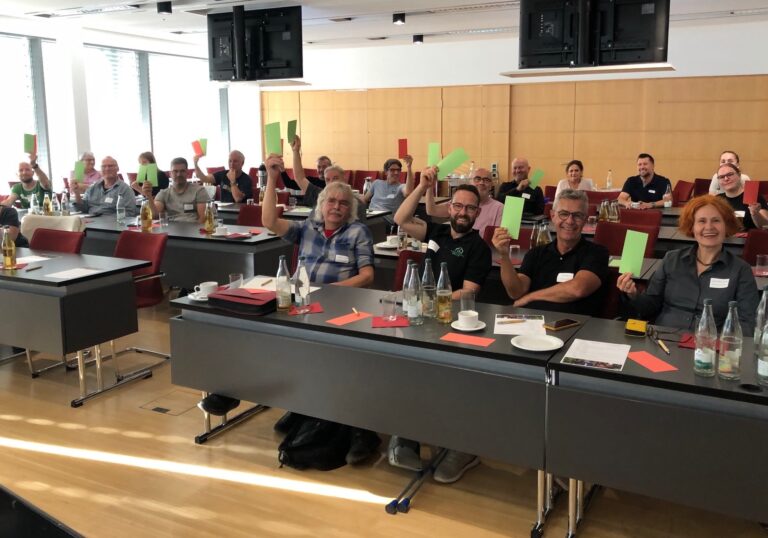 Group of adults in a classroom-style conference room, many holding up green and red cards during a discussion or vote, with bottles and papers on the desks.