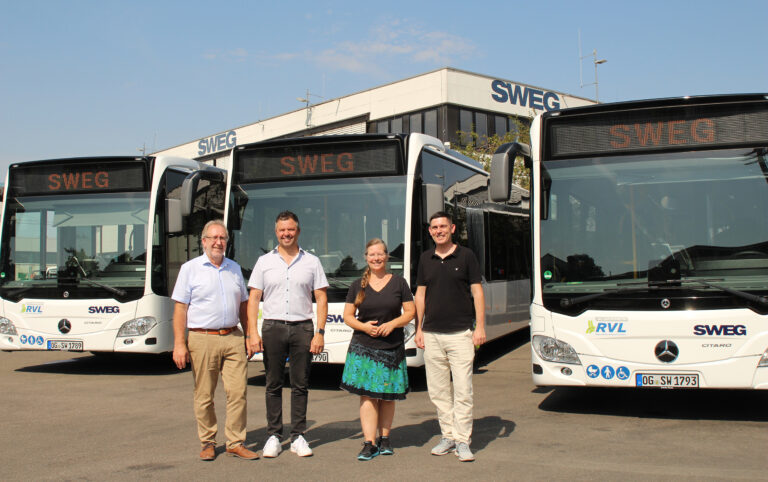 Four people stand in front of white buses labeled SWEG outside a building with a blue sky above.