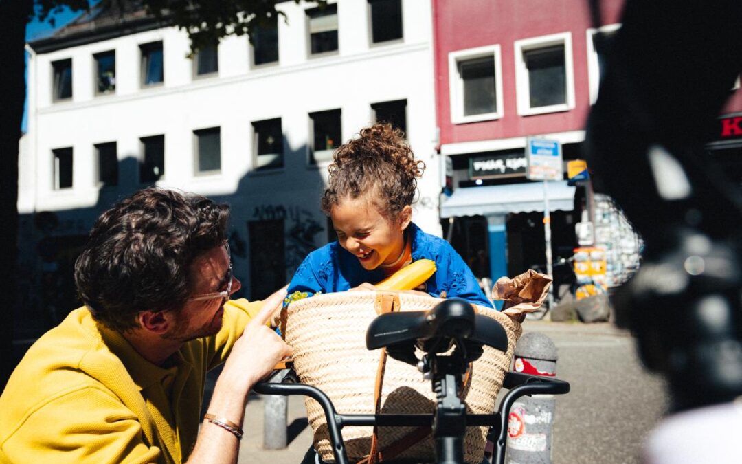 Man in a mustard jacket leans toward a smiling child in a wicker bike basket on a sunny city street.