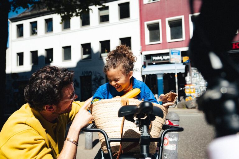 Man in a mustard jacket leans toward a smiling child in a wicker bike basket on a sunny city street.
