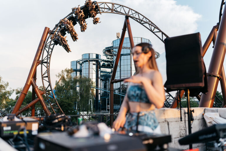 Riders on a steel roller coaster looping above, upside down mid-ride with park buildings in the background.