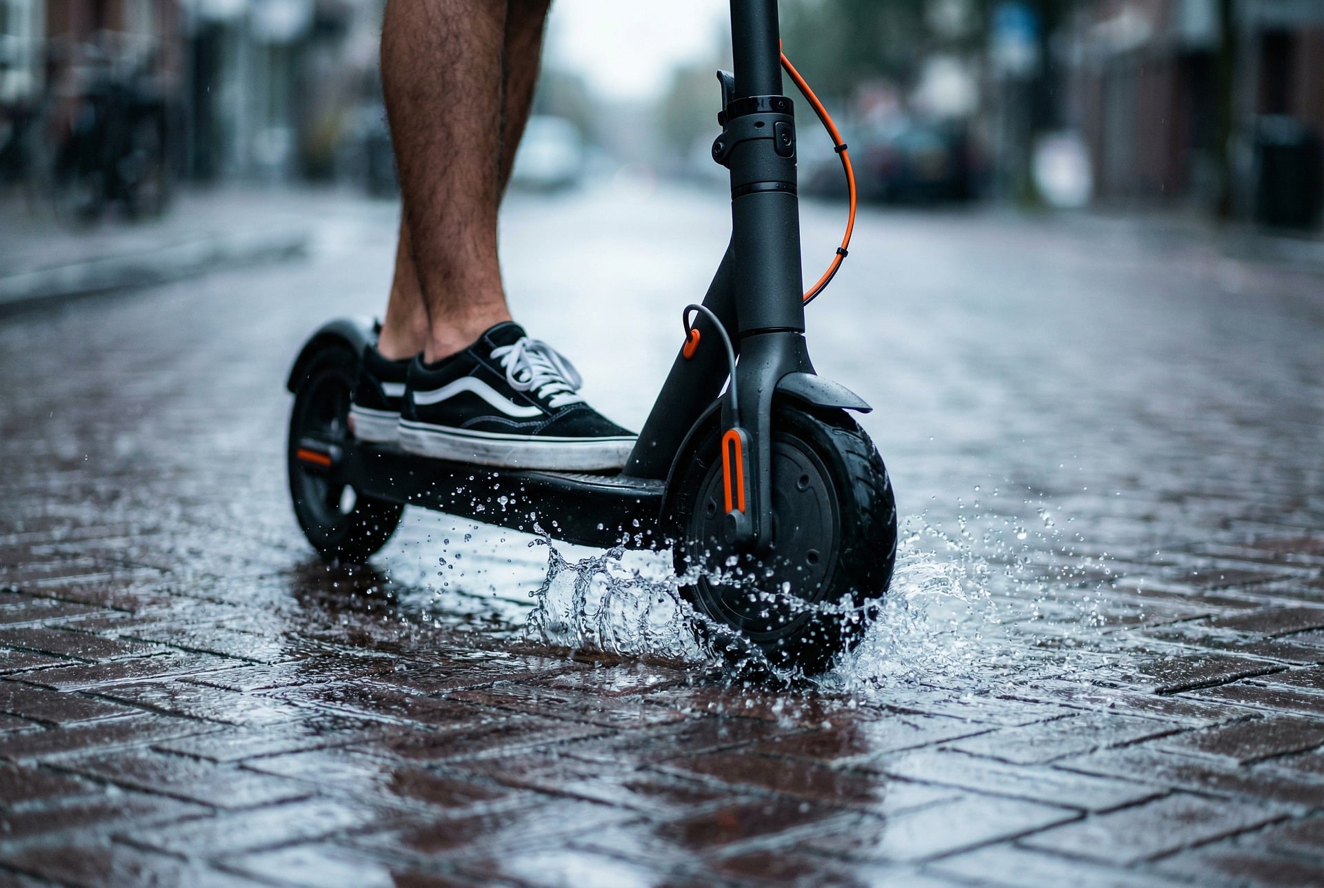 Person rides an electric scooter on a wet cobblestone street, splashing water with the front wheel.
