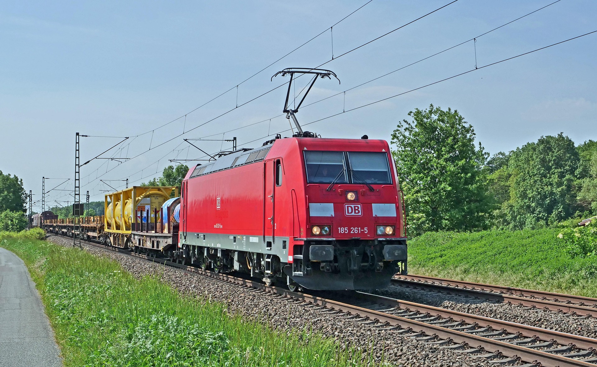 Red DB electric locomotive hauling freight cars along a track with yellow cylindrical tanks amid green fields and trees under a blue sky.