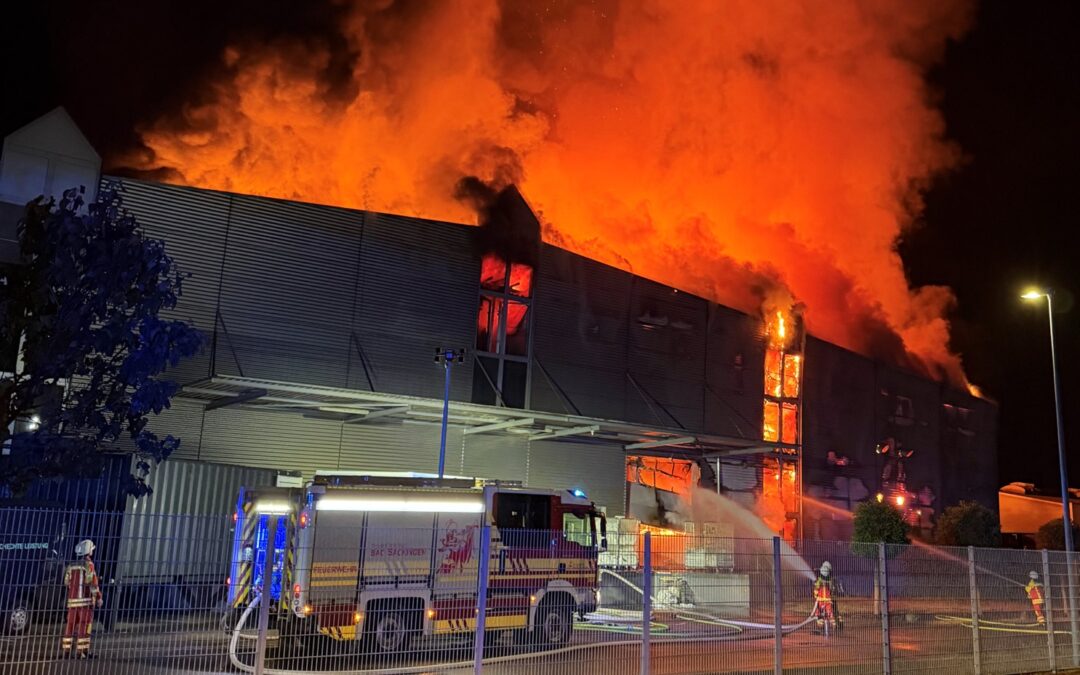 Nighttime industrial fire: a large warehouse is engulfed in orange flames with thick smoke as firefighters spray water from a fire truck by a fenced area.