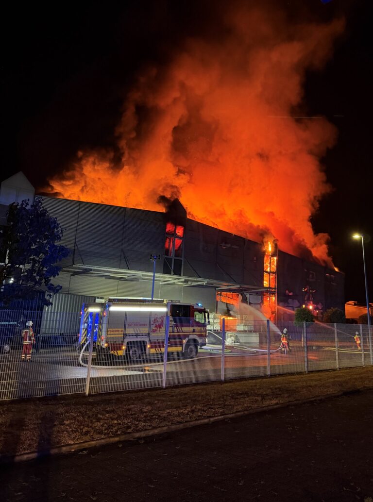 Nighttime industrial fire: a large warehouse is engulfed in orange flames with thick smoke as firefighters spray water from a fire truck by a fenced area.