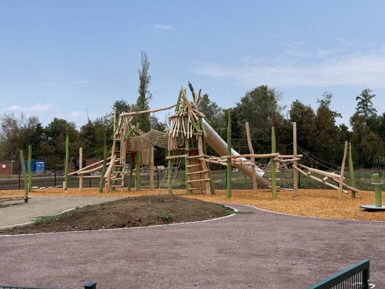 Wooden climbing structure with logs, tunnels and a slide in a playground, on wood-chip ground with trees in the background, under a blue sky.
