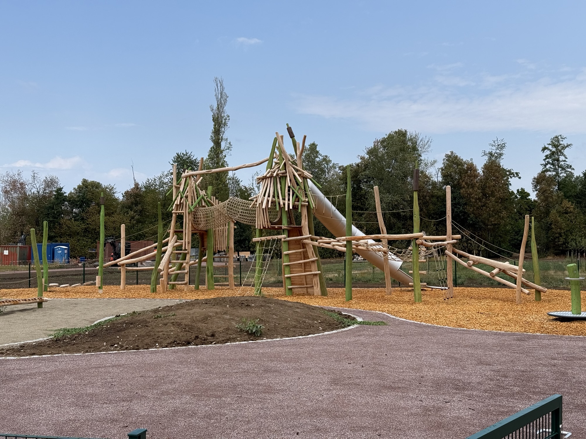 Wooden climbing structure with logs, tunnels and a slide in a playground, on wood-chip ground with trees in the background, under a blue sky.