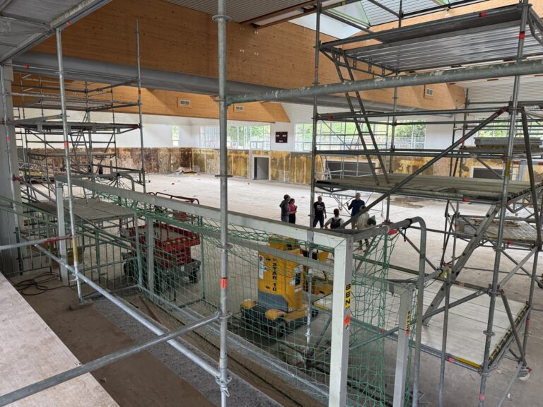 Construction site interior with metal scaffolding and wooden walls; a small group of workers in the background.