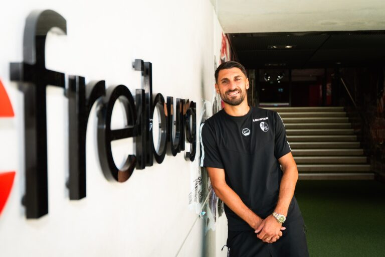 Man leaning against a wall with large 3D lettering in a hallway, wearing a black sports shirt and smiling at the camera.