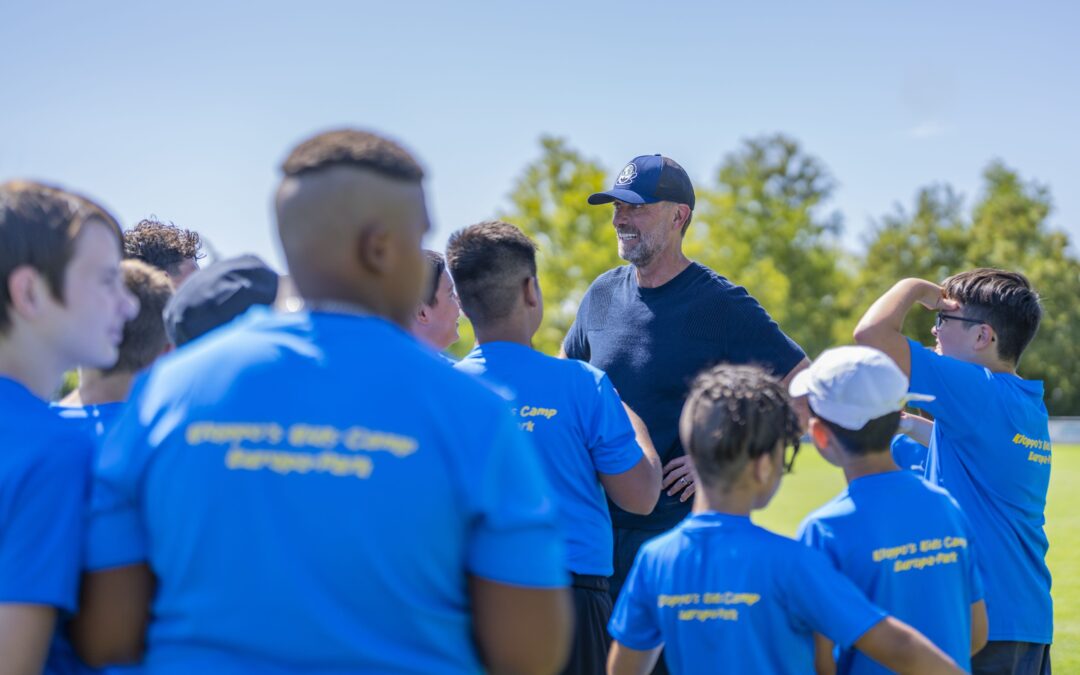Coach in a dark shirt talks to a group of athletes in blue shirts outdoors on a sunny day field.