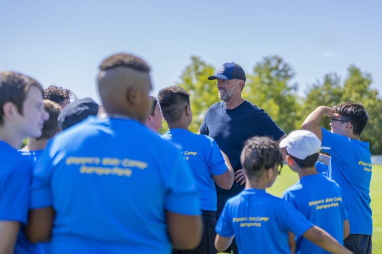 Coach in a dark shirt talks to a group of athletes in blue shirts outdoors on a sunny day field.