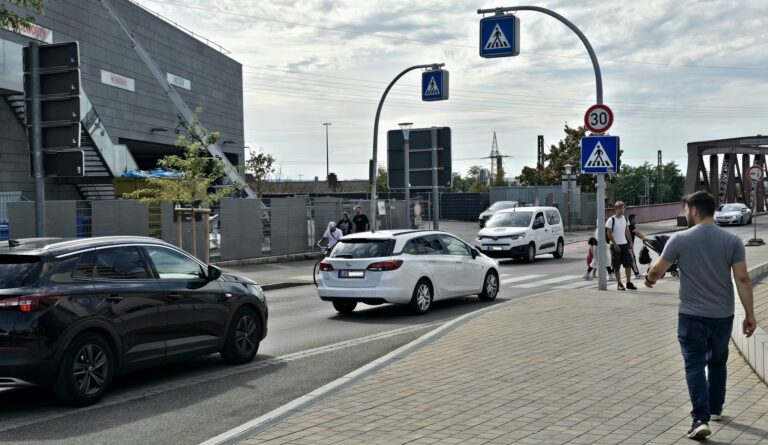 A busy city street with pedestrians crossing a crosswalk, several cars driving and a modern building under construction in the background.