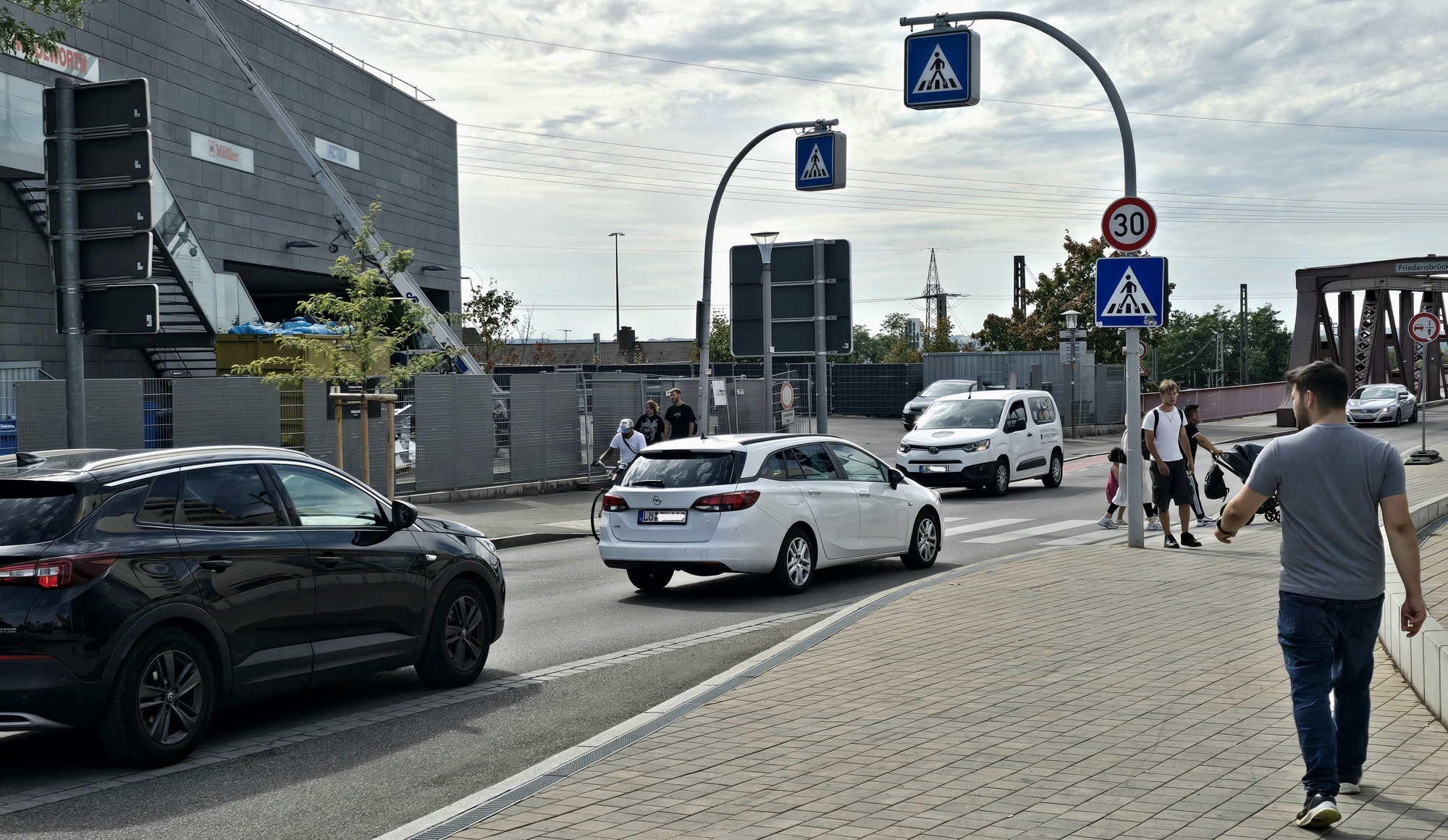 A busy city street with pedestrians crossing a crosswalk, several cars driving and a modern building under construction in the background.