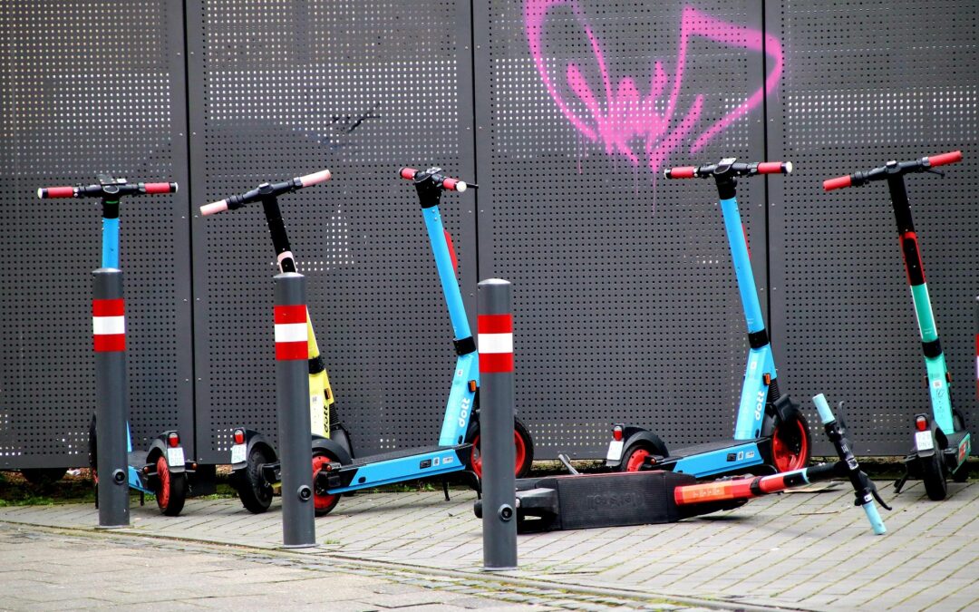 Blue and black kick scooters lined up at a bike rack in front of a perforated metal wall with pink graffiti.